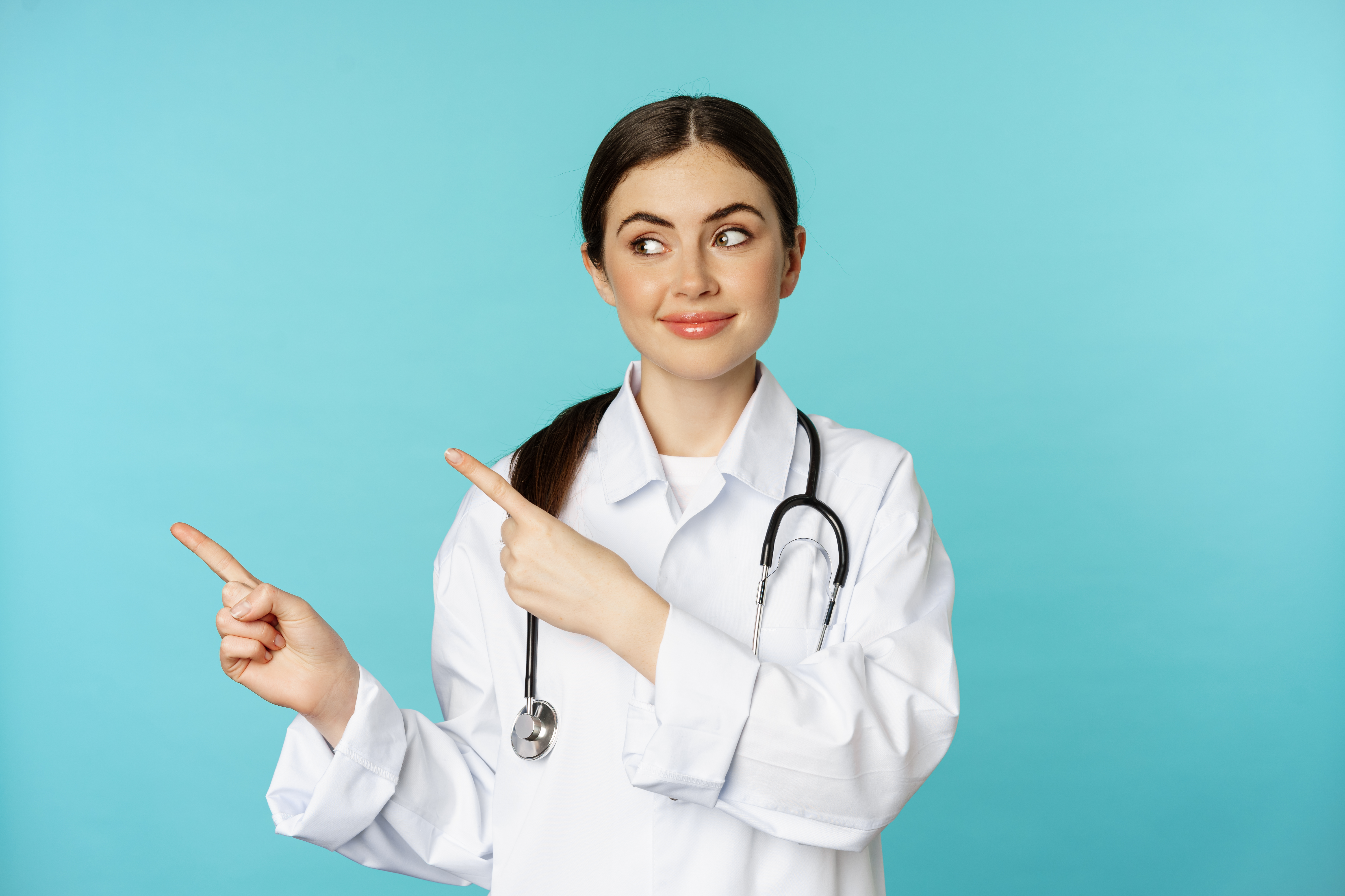 Portrait of smiling medical worker, girl doctor in white coat with stethoscope, pointing fingers left, showing medical clinic advertisement, torquoise background.